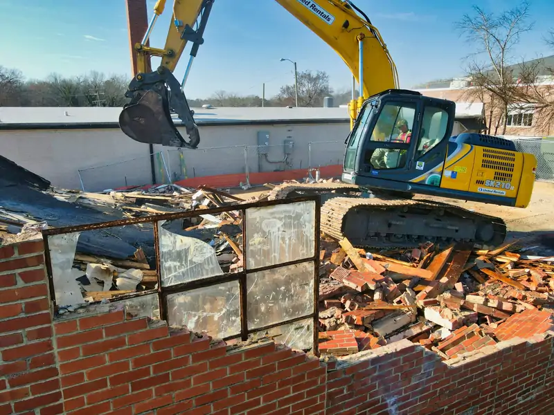 Excavator demolishing a brick building with the operator visible in the cab