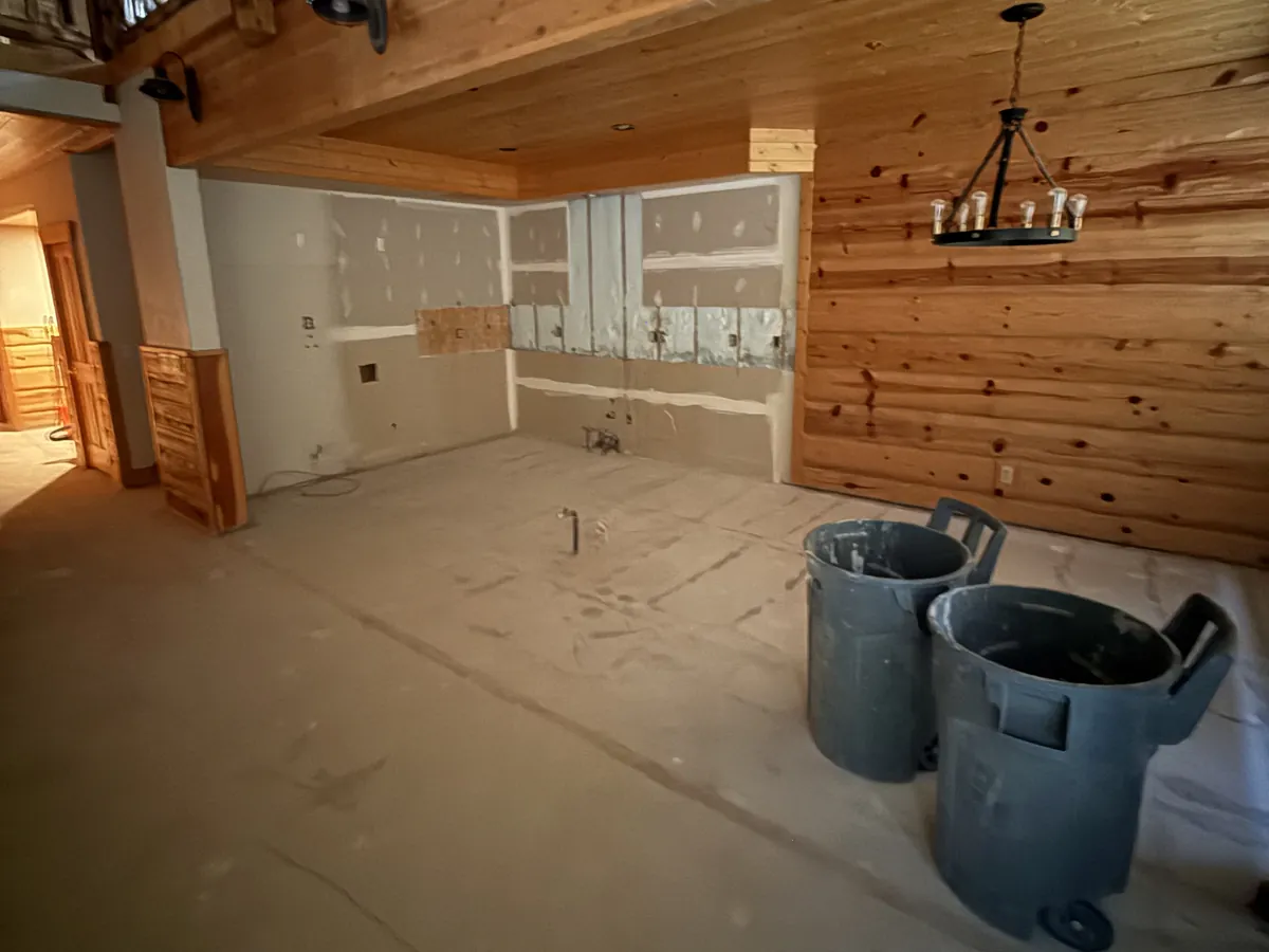 Wood-paneled cabin kitchen with the cabinets and counters stripped out and debris bins on the bare subfloor