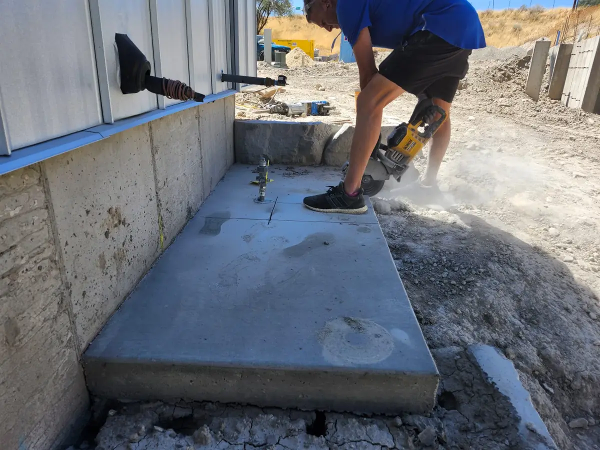 Worker saw-cutting a concrete slab along a foundation wall, dust coming off the blade