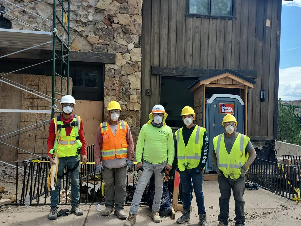 Five-person demolition crew in hard hats, hi-vis vests, and respirators in front of a house mid-demolition