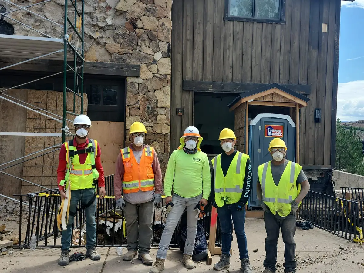 Five-person demolition crew in hard hats, hi-vis vests, and respirators in front of a house mid-demolition