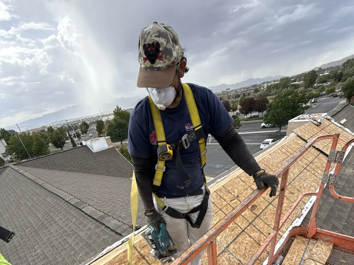 Worker in a fall-protection harness on a man lift at roof level, with the valley and mountains behind
